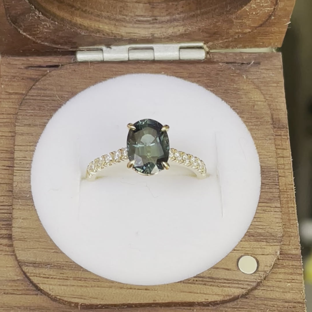 Oval Teal Sapphire ring set in four claws. The stone is beautifully complimented and by twelve 1.7mm round brilliant cut Diamonds and is set in 18ct yellow gold.  The Sapphire is 9 x 6.5mm 2.47ct, total diamond weight is approximately 0.24pts  The ring is Size L   Made with recycled metal in Sydney   Ethically sourced Gemstones