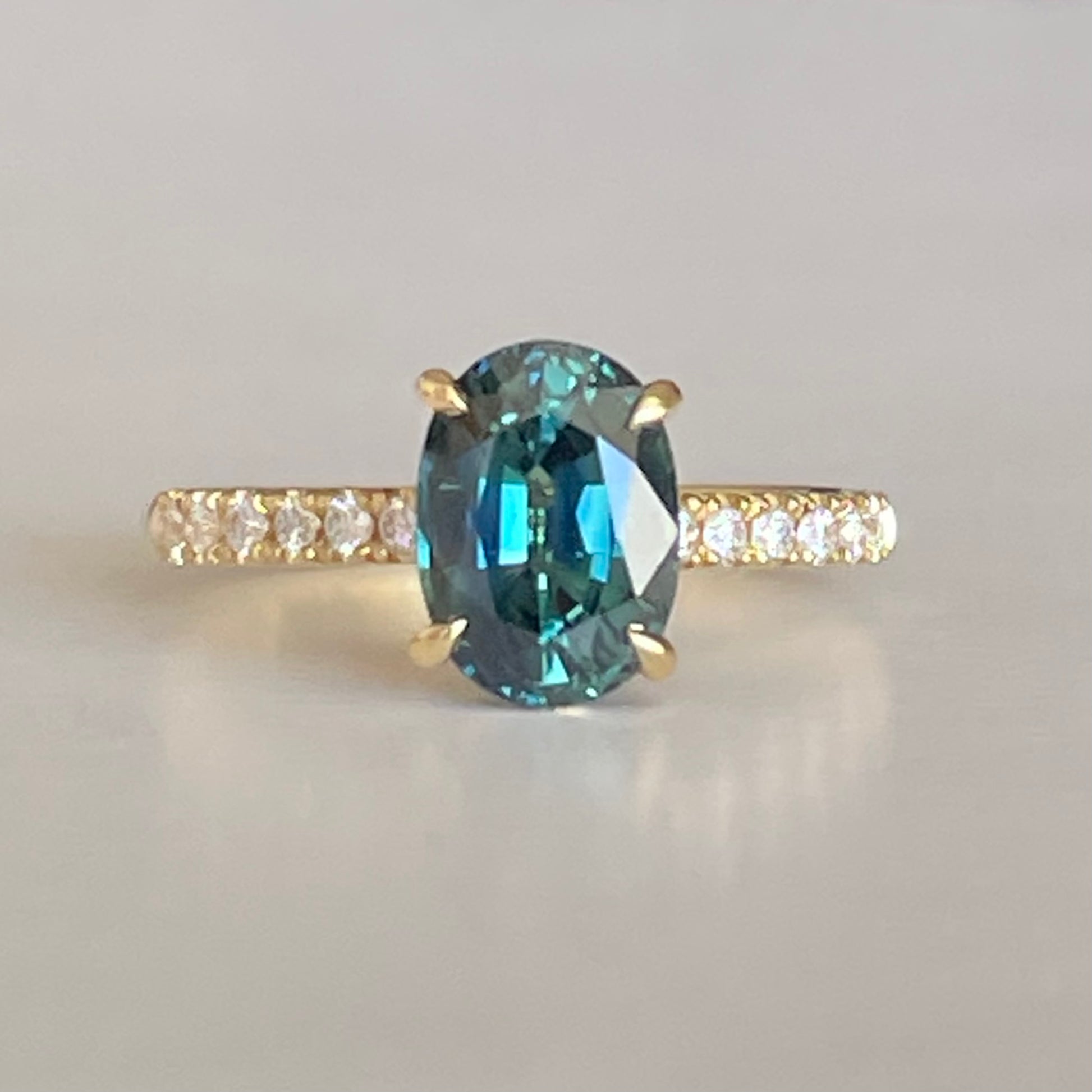 Oval Teal Sapphire ring set in four claws. The stone is beautifully complimented and by twelve 1.7mm round brilliant cut Diamonds and is set in 18ct yellow gold.  The Sapphire is 9 x 6.5mm 2.47ct, total diamond weight is approximately 0.24pts  The ring is Size L   Made with recycled metal in Sydney   Ethically sourced Gemstones