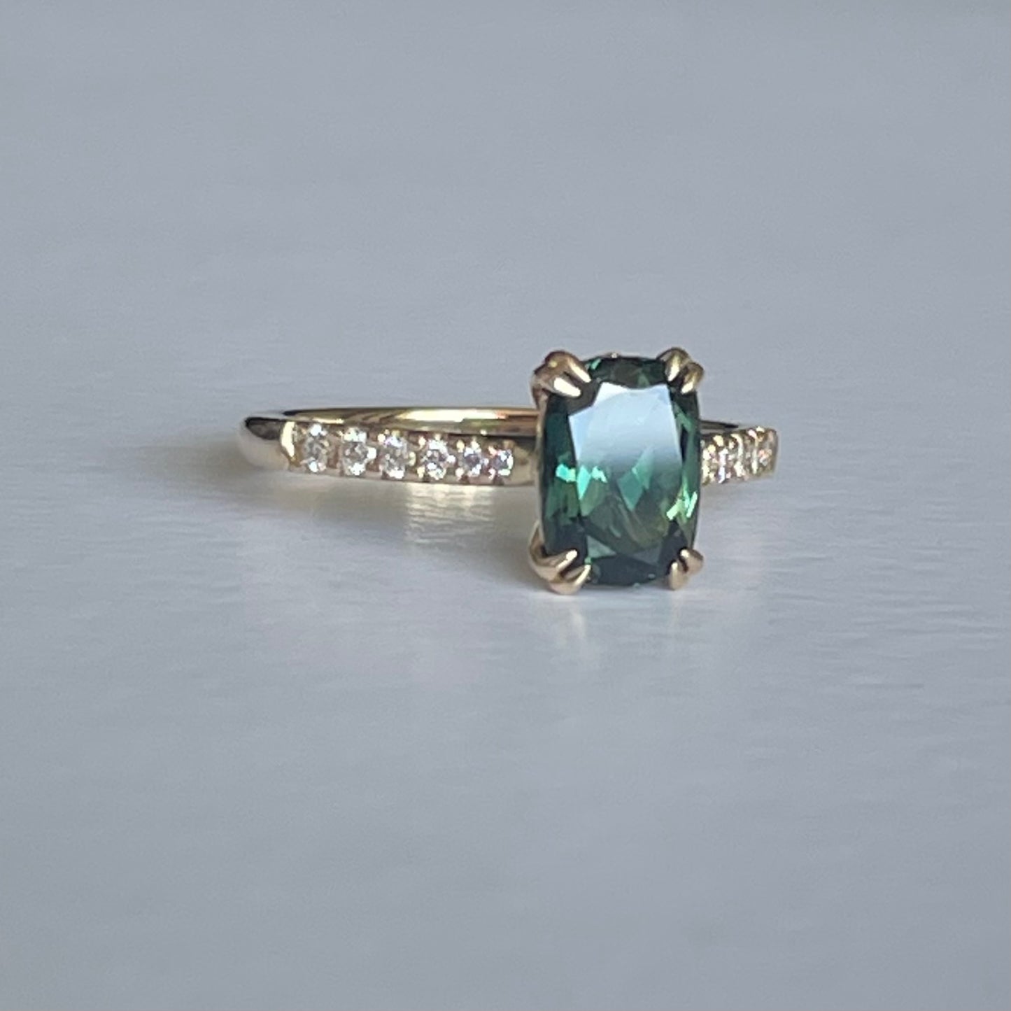 A Teal Green Australian Parti Sapphire from Rubyvale in QLD & is set with four double claws. The stone is beautifully complimented by 12 x Round Brilliant cut graduating diamonds total diamond weight 0.13pts and is set in 14ct Yellow gold. The Sapphire measures 7.9 x 5.9mm & is 1.75ct Available in Size M Made with recycled metal in Sydney Ethically sourced Gemstones