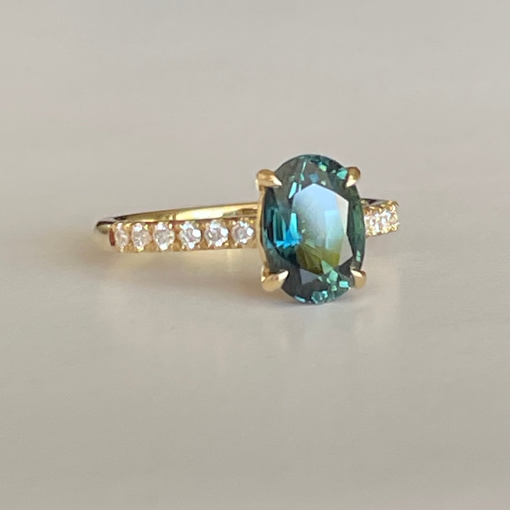 Oval Teal Sapphire ring set in four claws. The stone is beautifully complimented and by twelve 1.7mm round brilliant cut Diamonds and is set in 18ct yellow gold.  The Sapphire is 9 x 6.5mm 2.47ct, total diamond weight is approximately 0.24pts  The ring is Size L   Made with recycled metal in Sydney   Ethically sourced Gemstones