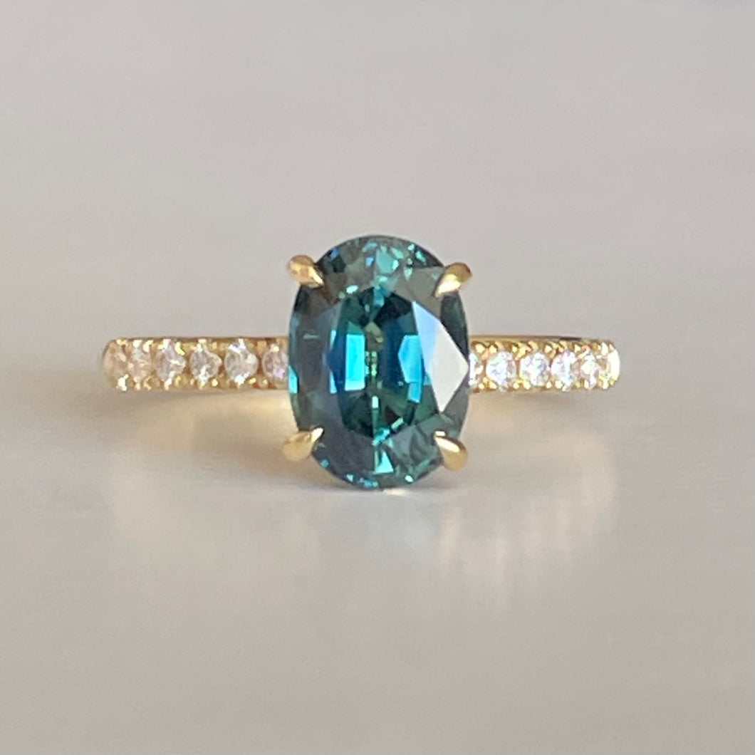 Oval Teal Sapphire ring set in four claws. The stone is beautifully complimented and by twelve 1.7mm round brilliant cut Diamonds and is set in 18ct yellow gold.  The Sapphire is 9 x 6.5mm 2.47ct, total diamond weight is approximately 0.24pts  The ring is Size L   Made with recycled metal in Sydney   Ethically sourced Gemstones 