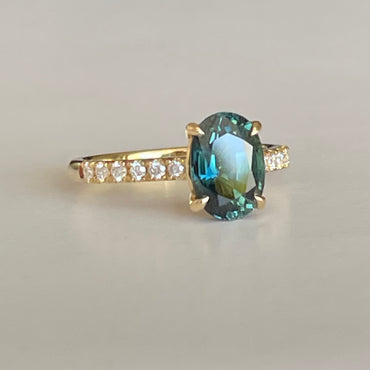 Oval Teal Sapphire ring set in four claws. The stone is beautifully complimented and by twelve 1.7mm round brilliant cut Diamonds and is set in 18ct yellow gold.  The Sapphire is 9 x 6.5mm 2.47ct, total diamond weight is approximately 0.24pts  The ring is Size L   Made with recycled metal in Sydney   Ethically sourced Gemstones 